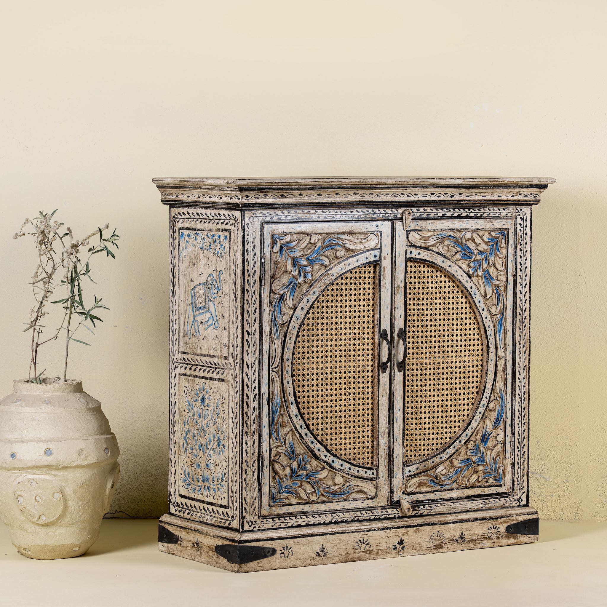Side view of a hand-made, hand-carved cupboard made of mango wood with blue floral designs, elephant motifs and rattan inserts.