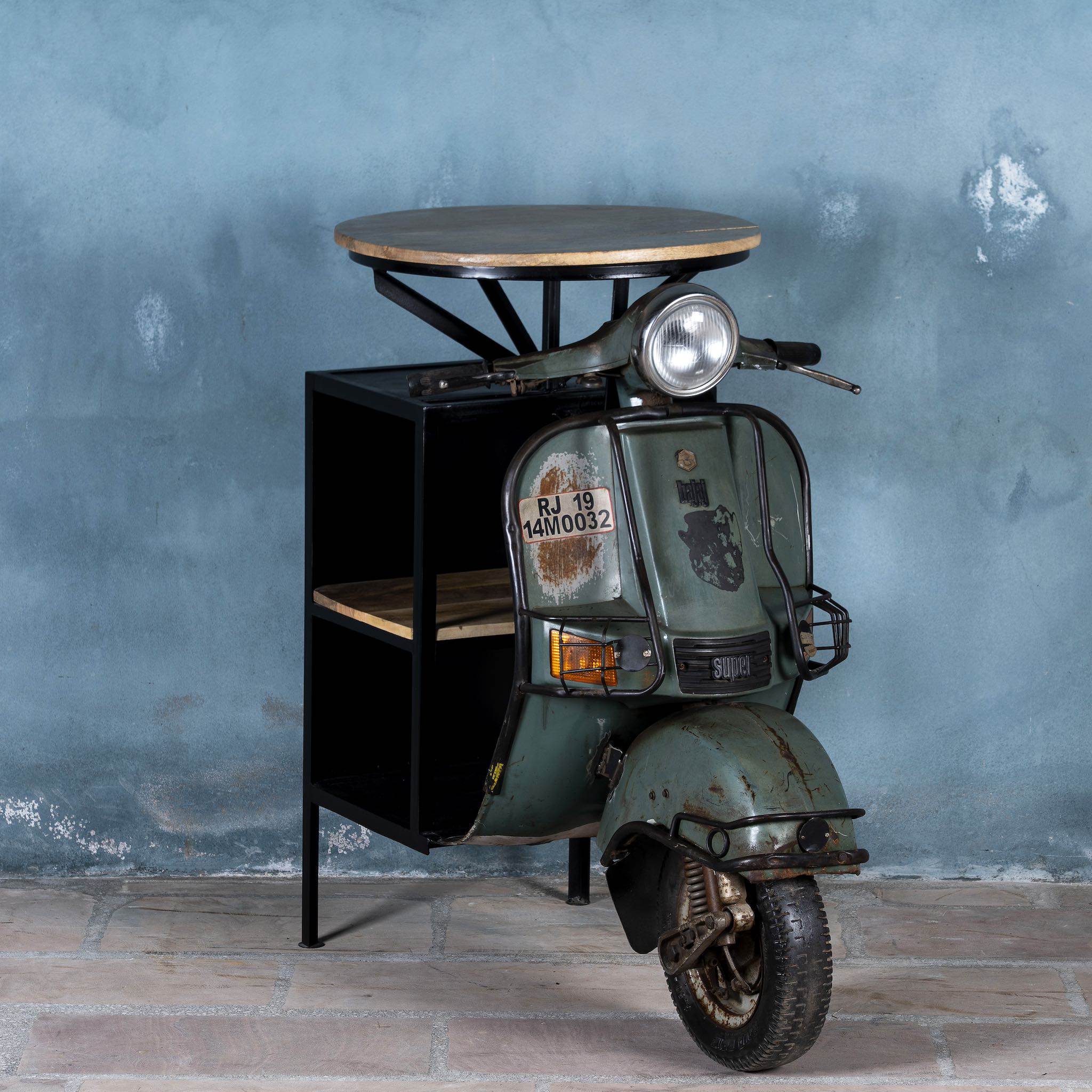 Side angle image of a short bar table crafted from the front half of an original Bajaj scooter, featuring a round wooden top, metal frame, and rustic industrial finish.