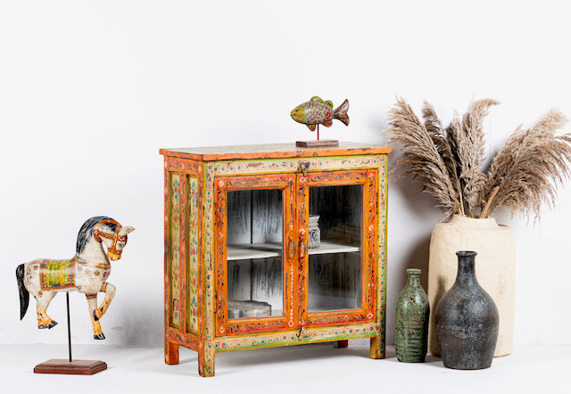Side angle image of a hand-painted teak wood display cabinet with glass doors and vibrant Indian motifs – sustainable furniture with vintage charm and artisanal detail.