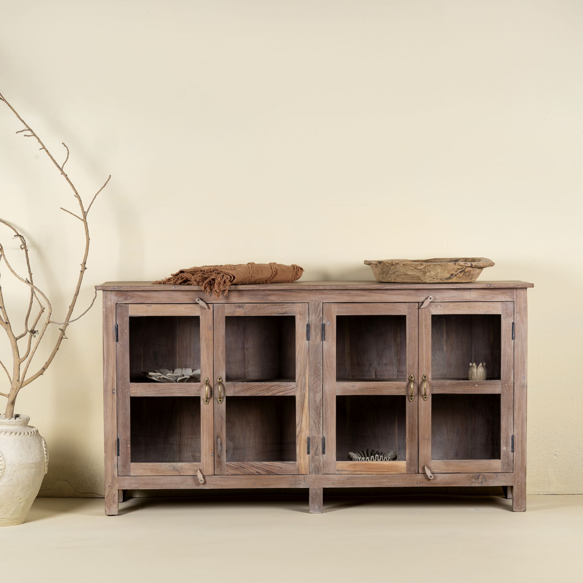 Front view image of a rustic showcase cabinet made from recycled teak wood, featuring four glass-paneled doors with brass handles and a lightly weathered finish, offering a warm, natural look ideal for displaying decor or tableware.