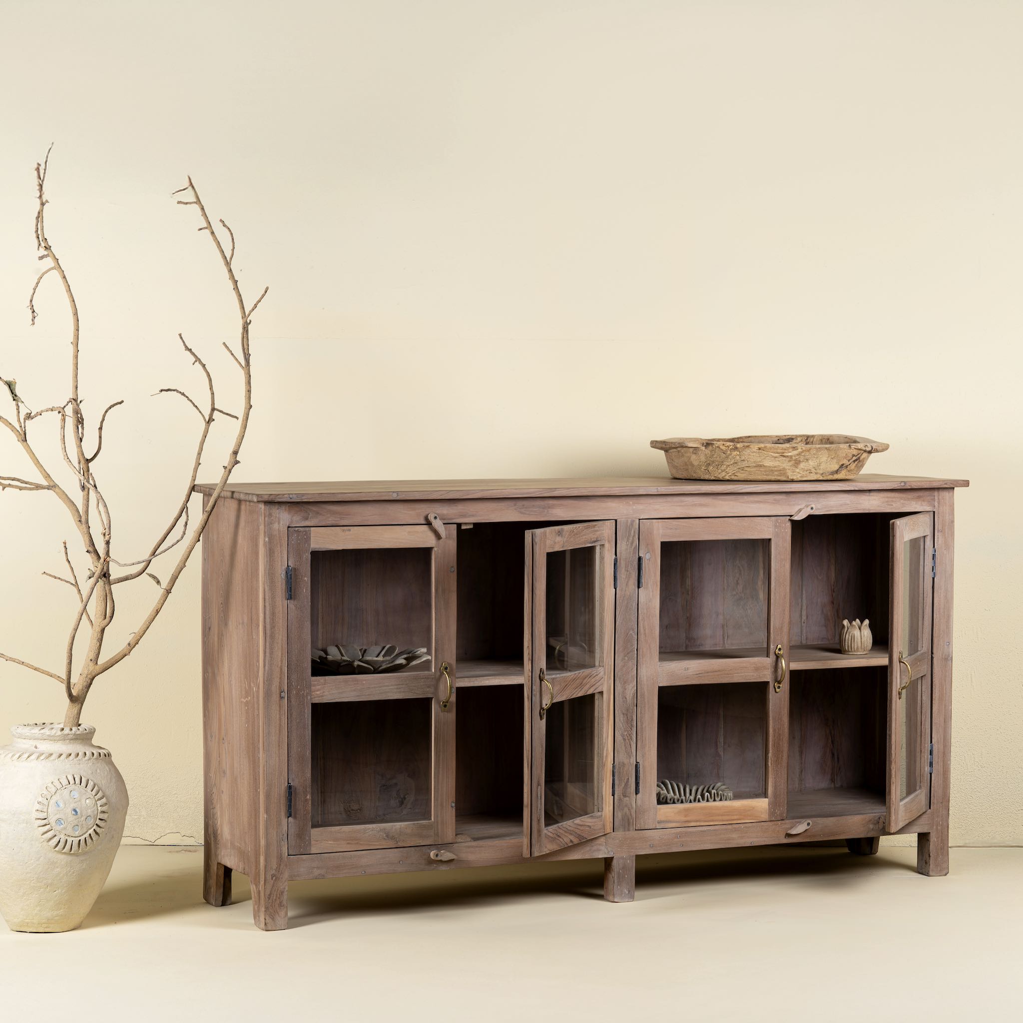 Side angle with open doors image of a rustic showcase cabinet made from recycled teak wood, featuring four glass-paneled doors with brass handles and a lightly weathered finish, offering a warm, natural look ideal for displaying decor or tableware.