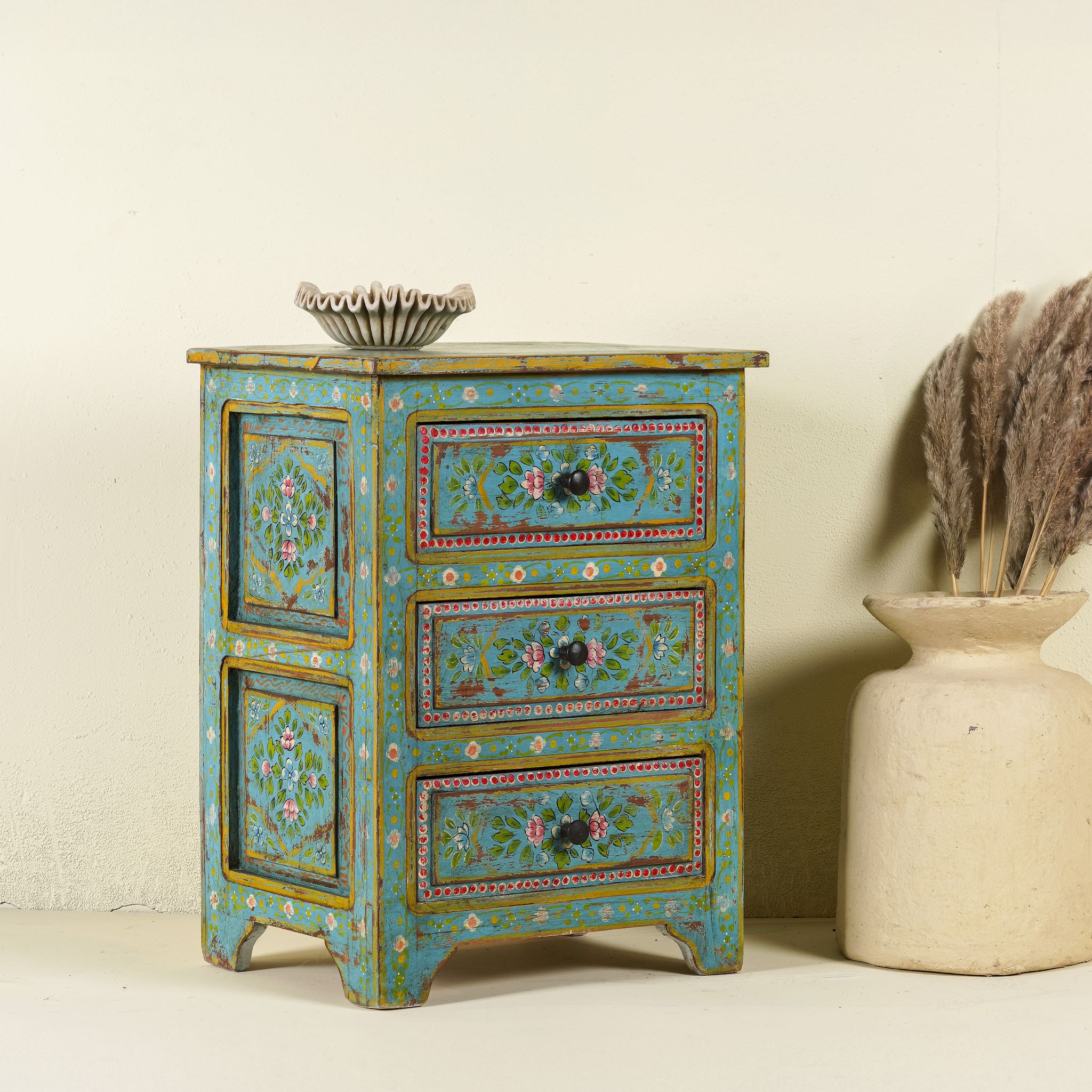 Side angle image of a hand-painted reclaimed wood nightstand with vibrant colours and floral patterns, featuring three drawers, placed next to a vase with dried plants.