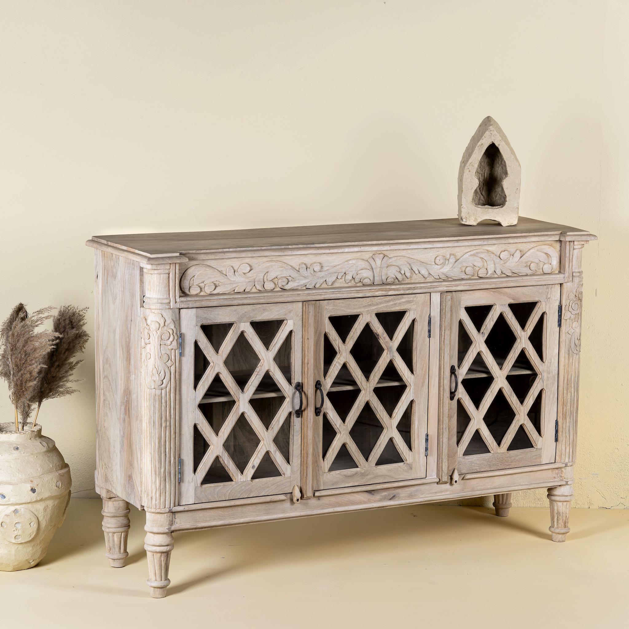 Side angle image of a rustic sideboard made of Mango wood with hand-carved details and glass panelled doors featuring a diamond lattice design.