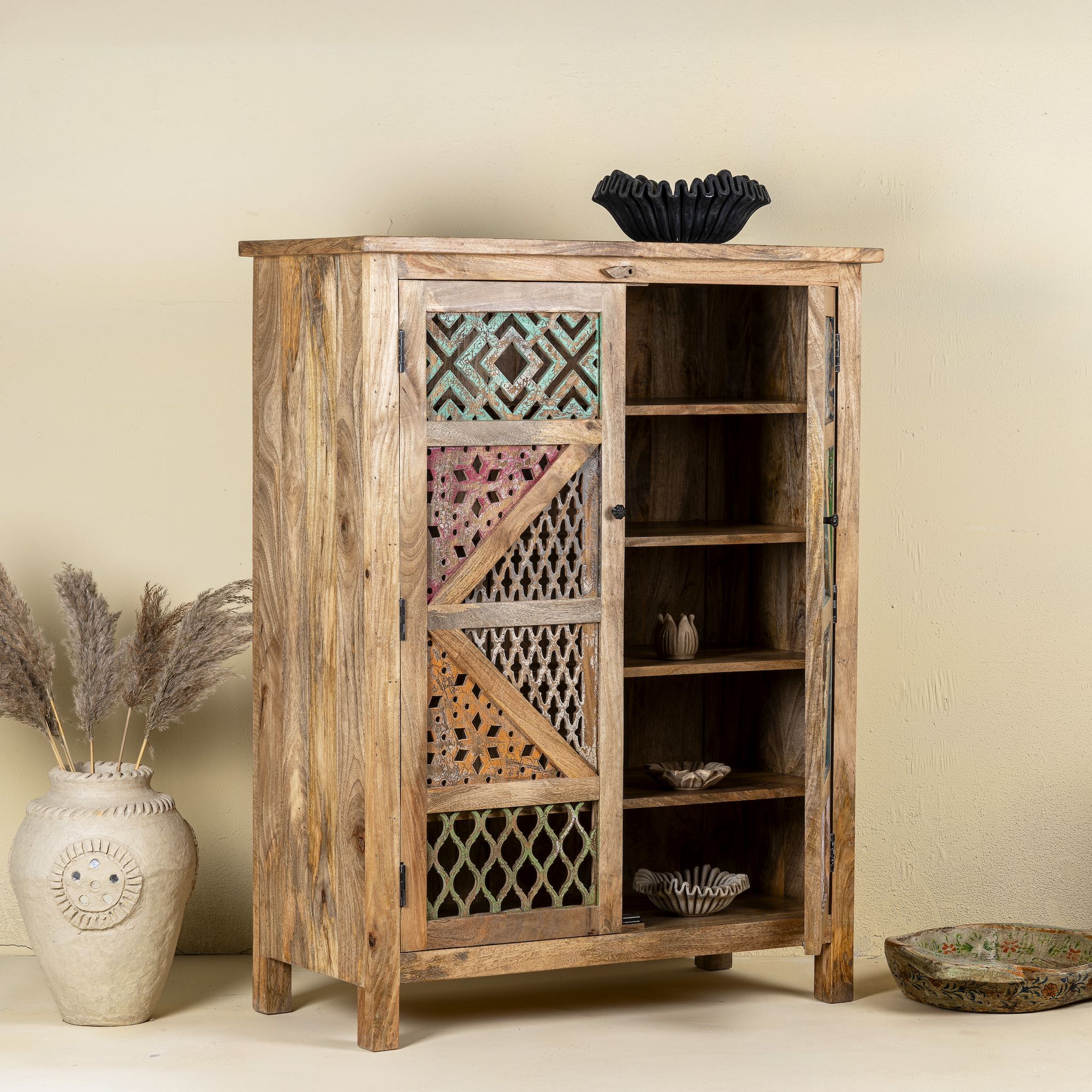 Side angle image with open door of a handmade mango wood cabinet with carved floral and geometric lattice doors, distressed colour accents; artisanal furniture perfect for boho style interiors, 100 × 45 × 130 cm.