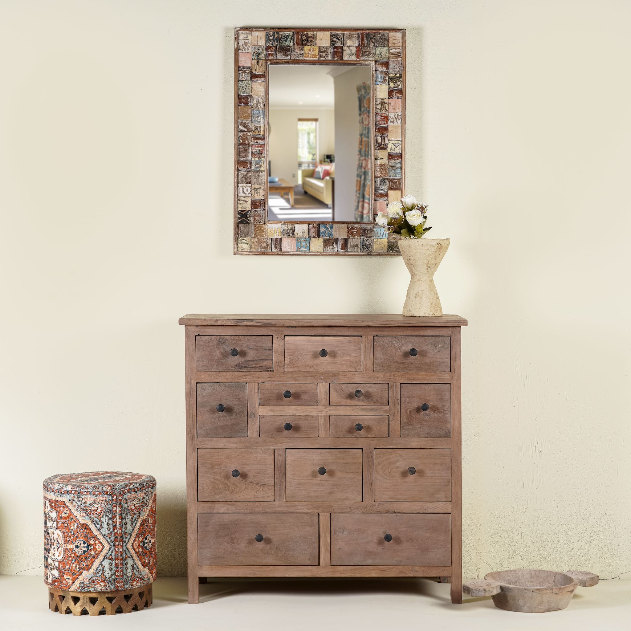 A handcrafted teak wood chest of drawers with a mirror above it, decorative items on top, and a bohemian style pattern on the drawer front.