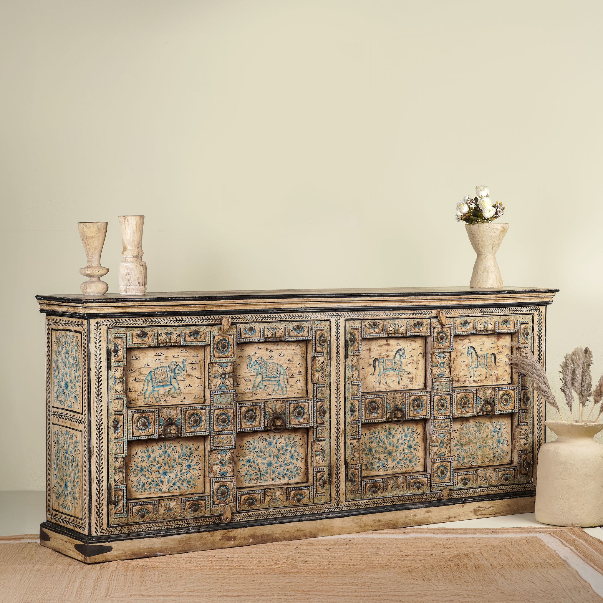 Side angle image of a vintage sideboard made from teak wood, hand-painted with traditional Indian motifs including elephants, camels, horses, and floral patterns in muted teal and earthy tones, featuring detailed borders and a distressed finish.