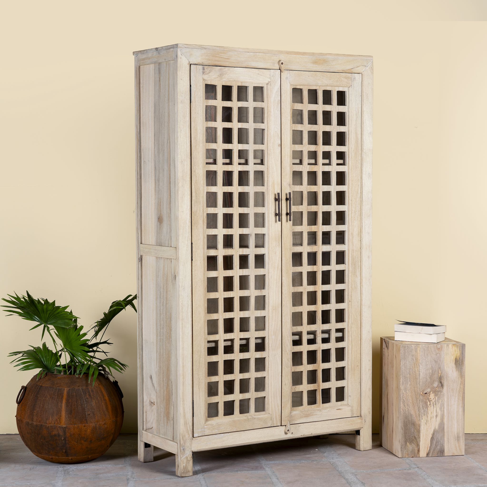 Side angle image of a sandblasted mango wood showcase with two glass-panelled grid doors and four interior compartments, finished in a light sun-washed tone, ideal as living room furniture.