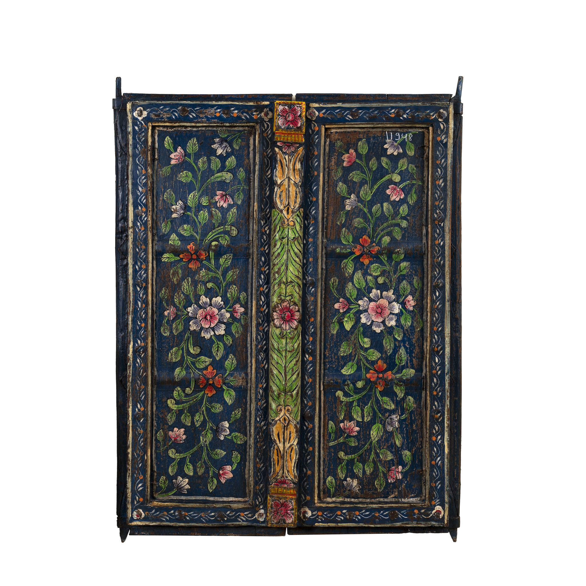 White background image of a hand-painted wall panel made from an old teak wood window frame, adorned with detailed floral motifs in shades of pink, red, green, and white against a deep blue background, framed by intricate carvings and colorful decorative accents.
