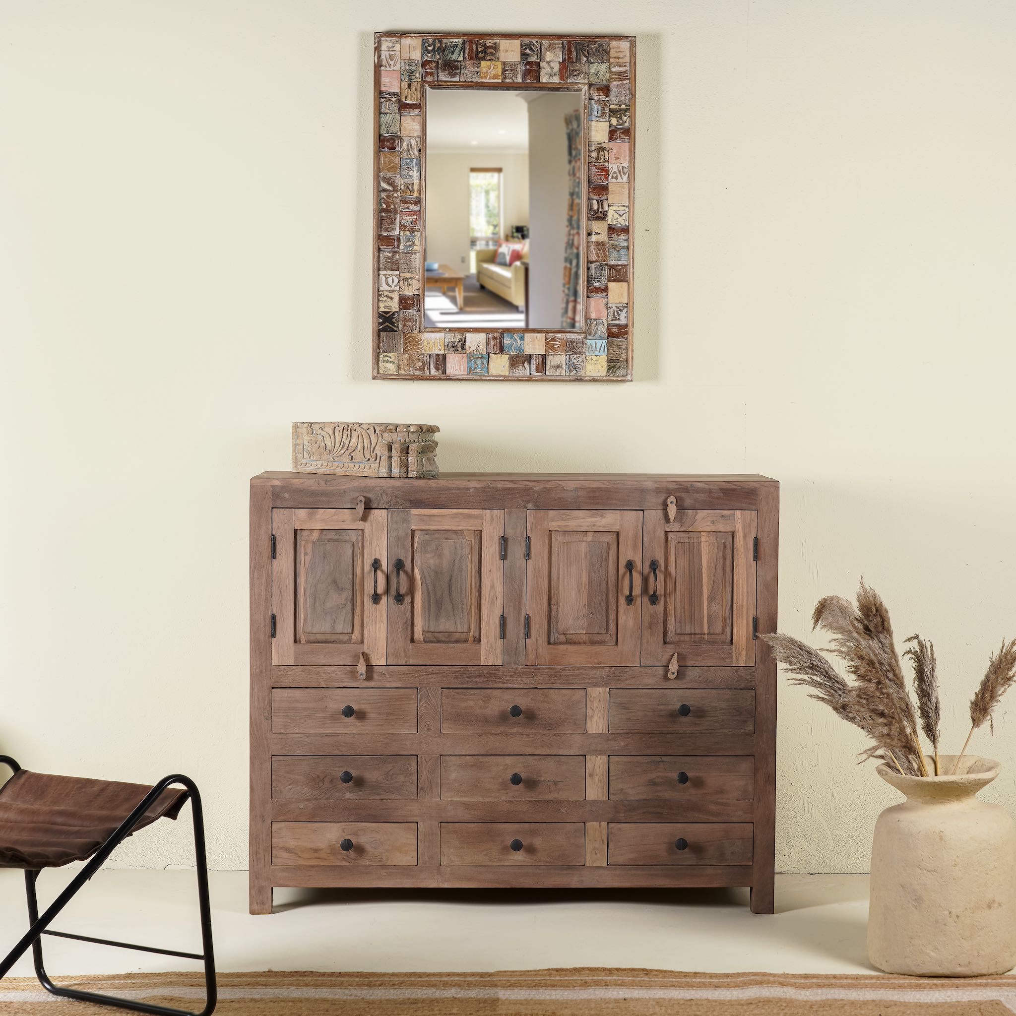 An elegant teak wood sideboard with multiple drawers and cupboards, featuring a brown finish. It is placed against a wall with a decorative mirror above it, and there is a vase with dried plants to the side.