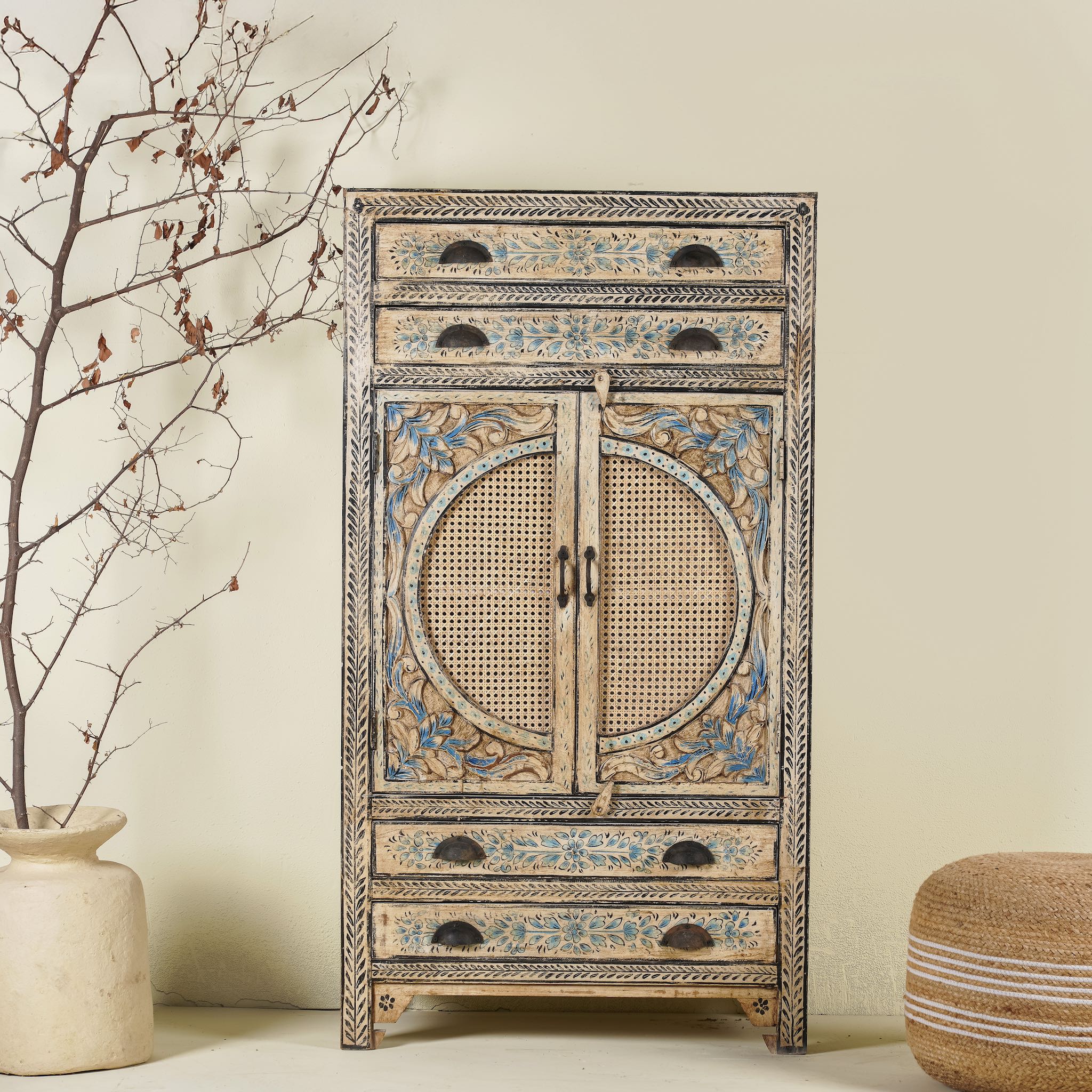 A hand-painted mango wood cabinet with rattan inserts, featuring floral patterns in blue and earthy tones, alongside a branch vase and a woven basket.