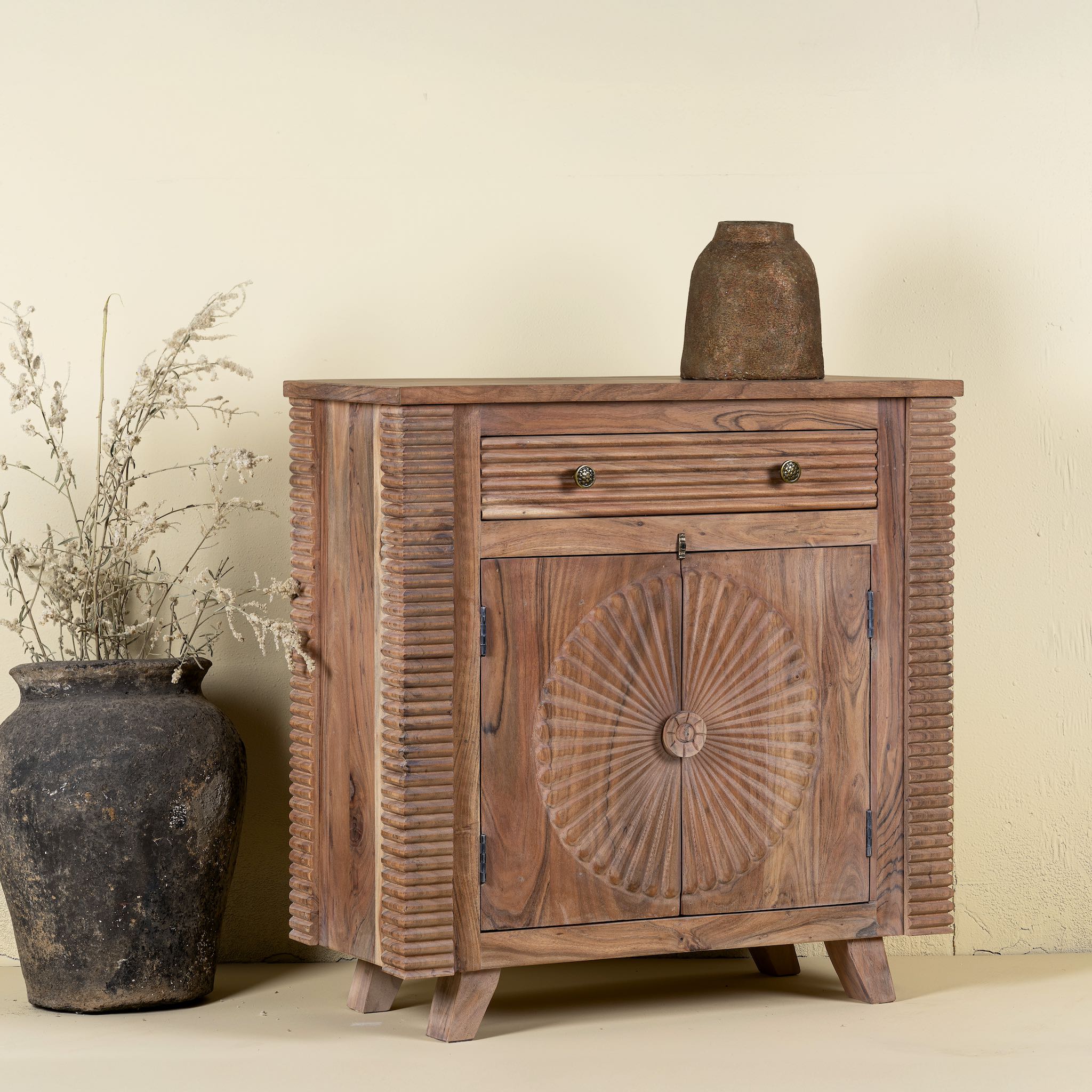 Side angle image of a acacia wood cabinet with hand-carved sunburst doors, grooved details, and one drawer, 90 × 40 × 90 cm.