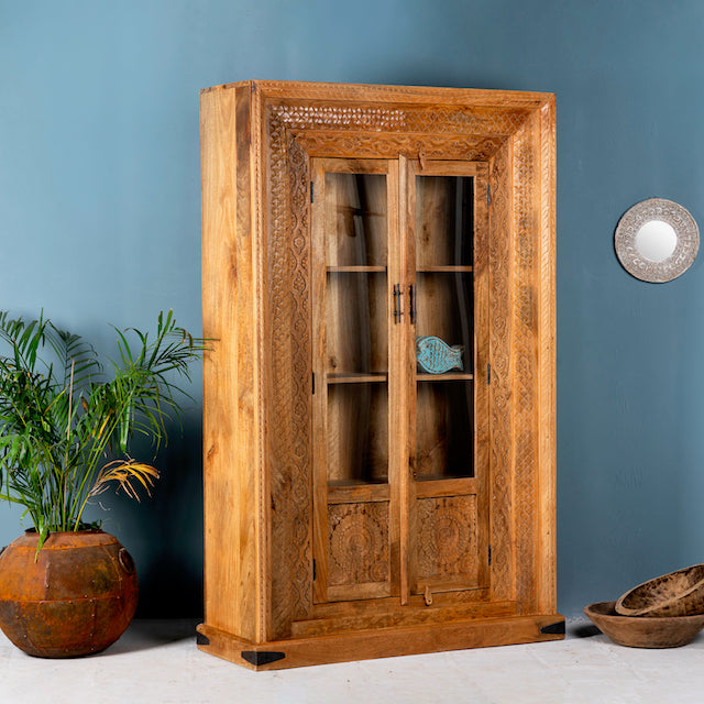 Side angle image of a hand carved display cabinet made from natural mango wood with floral detailing and glass-panelled doors; 180 × 110 × 40 cm.
