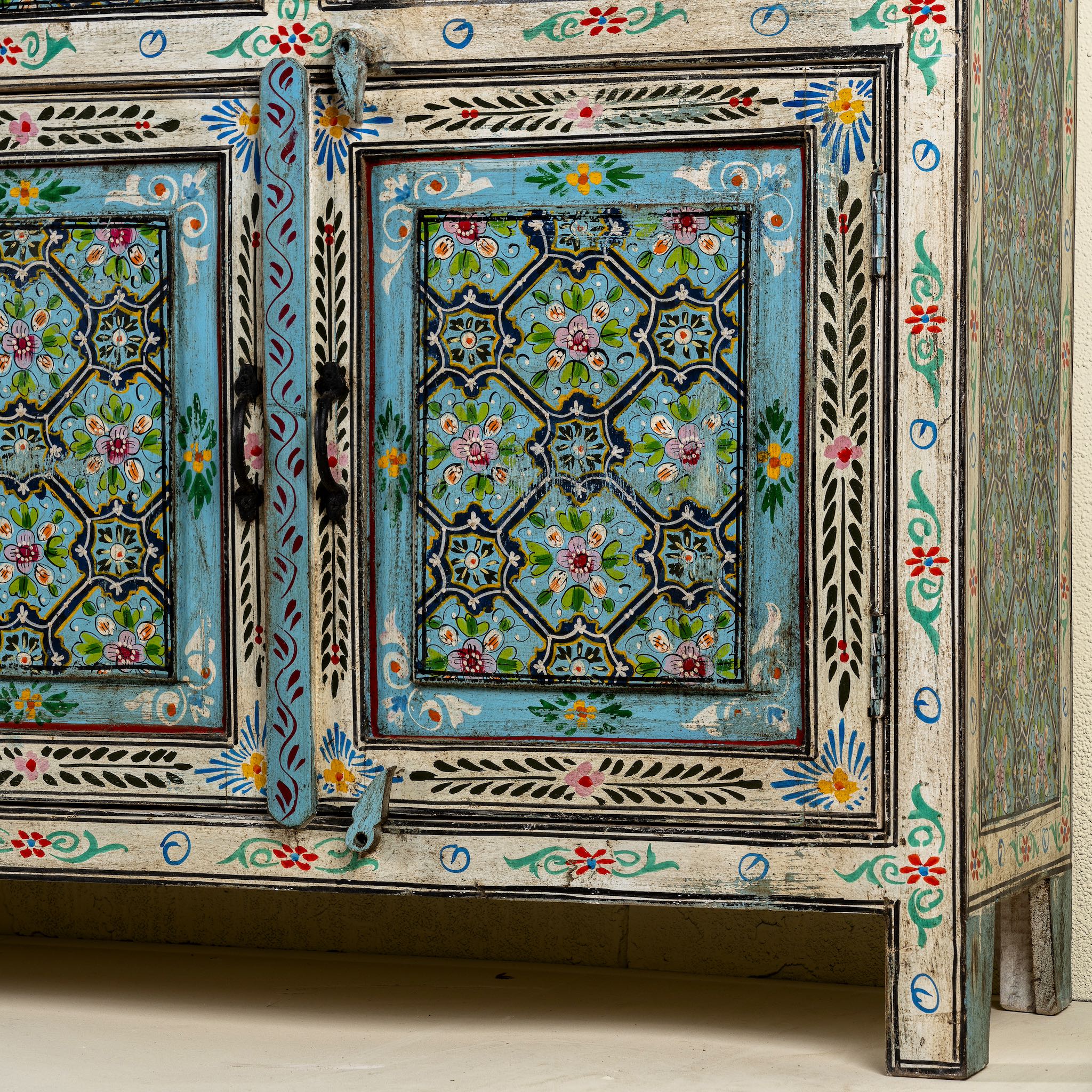 Close up image of the leg part of a hand-painted reclaimed wood sideboard with floral and geometric patterns in vibrant hues. The sideboard has multiple drawers and cabinet doors, and is placed in a room with a vase on top and a chair to the side.