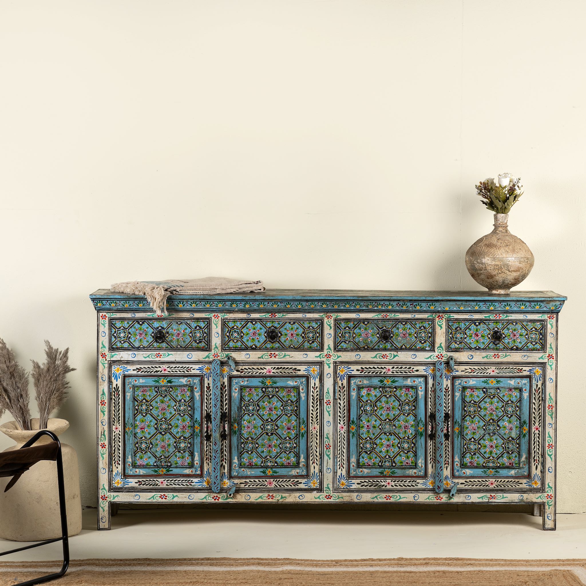 A hand-painted reclaimed wood sideboard with floral and geometric patterns in vibrant hues. The sideboard has multiple drawers and cabinet doors, and is placed in a room with a vase on top and a chair to the side.