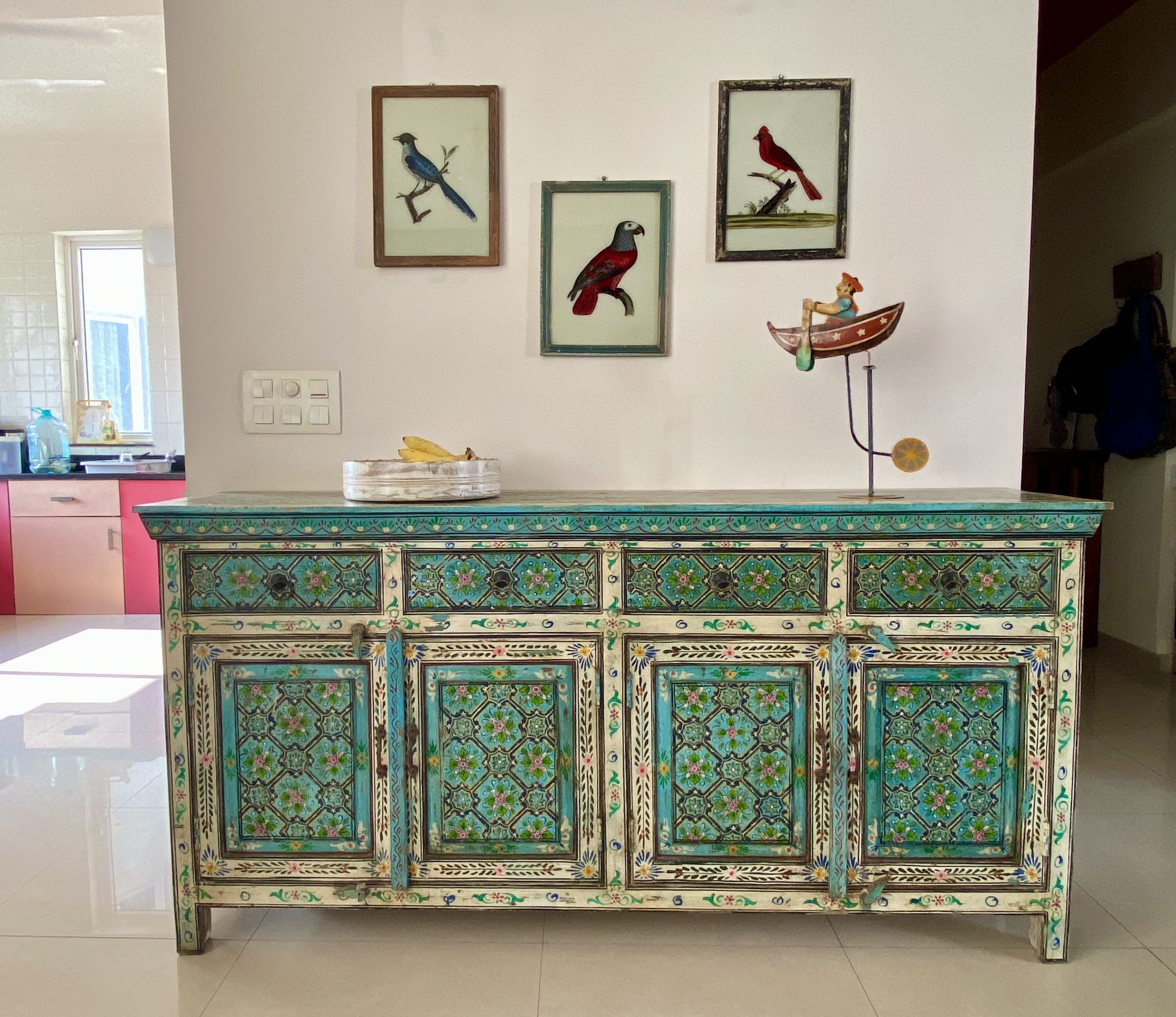 Lifestyle image of a hand-painted reclaimed wood sideboard with floral and geometric patterns in vibrant hues. The sideboard has multiple drawers and cabinet doors, and is placed in a room with a vase on top and a chair to the side.