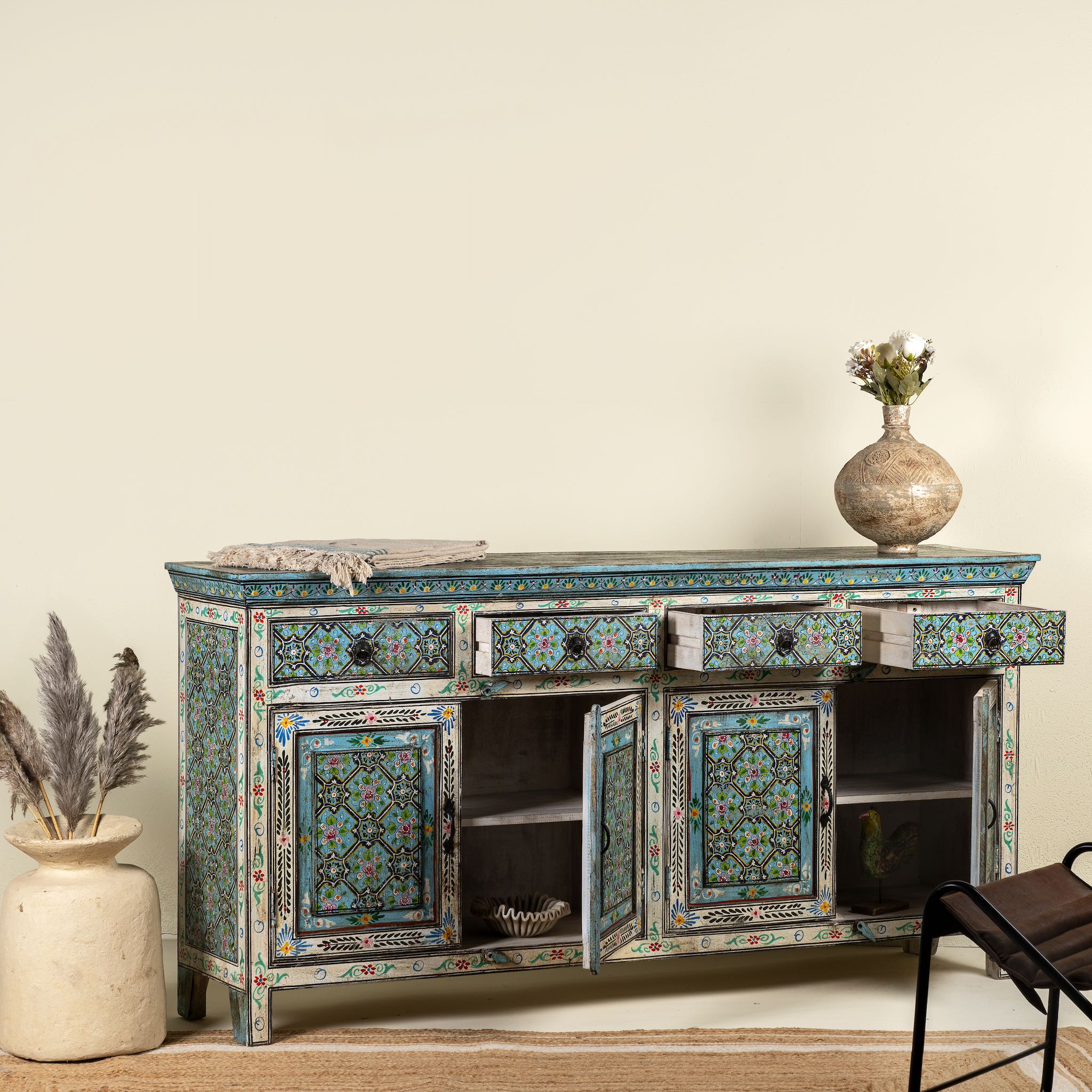 Open drawer image of a hand-painted reclaimed wood sideboard with floral and geometric patterns in vibrant hues. The sideboard has multiple drawers and cabinet doors, and is placed in a room with a vase on top and a chair to the side.