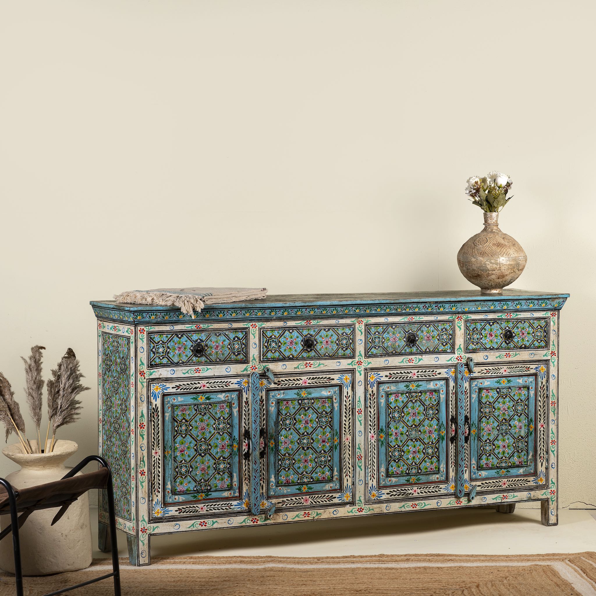 Side angle image of a hand-painted reclaimed wood sideboard with floral and geometric patterns in vibrant hues. The sideboard has multiple drawers and cabinet doors, and is placed in a room with a vase on top and a chair to the side.