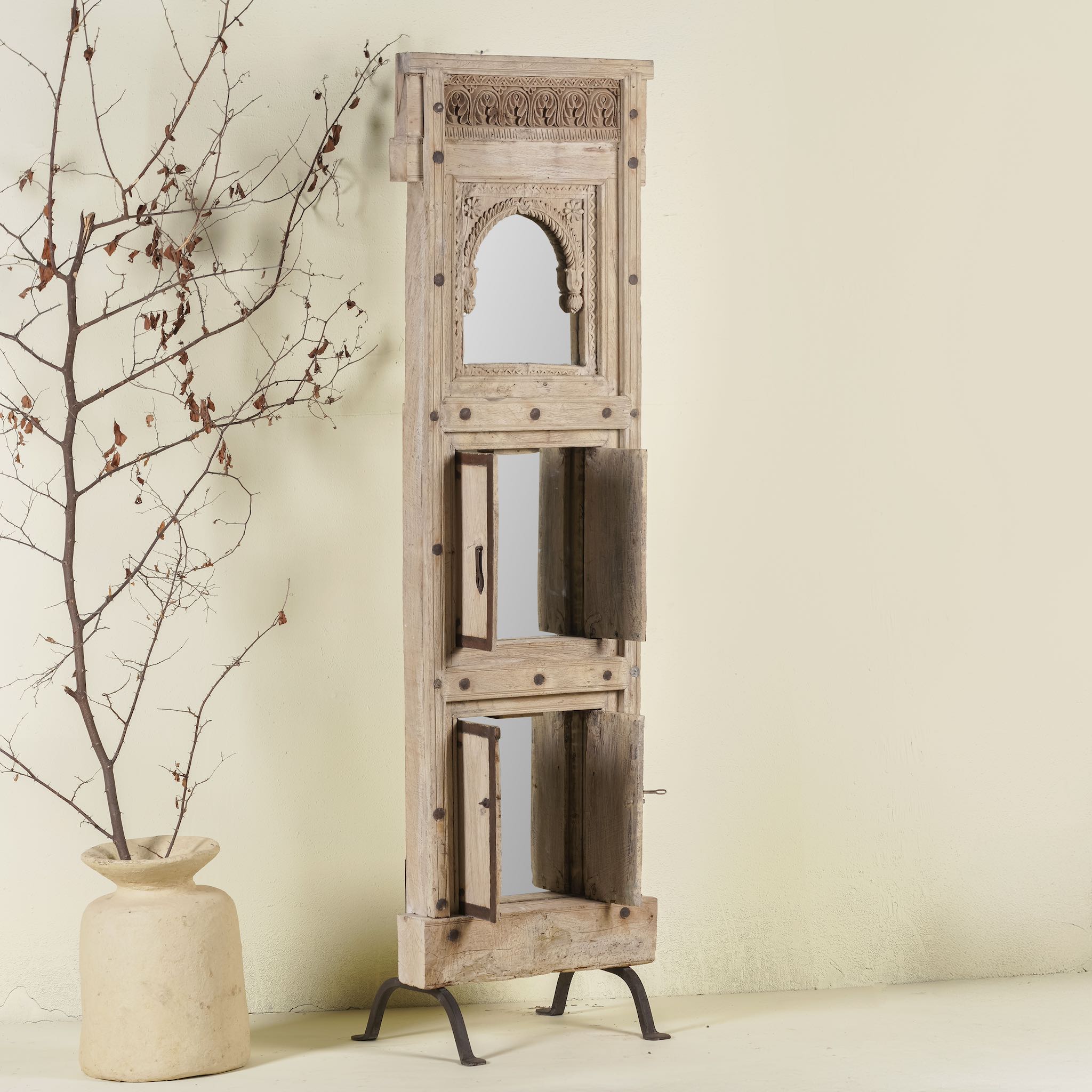 Side angle with open shutter of a mirror stand crafted from reclaimed teak wood with intricate carved detailing and two storage cabinets, set on a sturdy iron base, blending rustic charm with vintage Indian architectural elements.