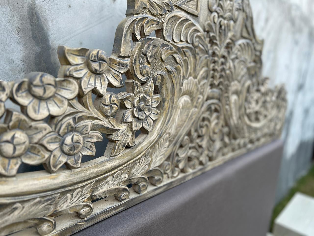Close up image of a hand-carved vintage bed Samar made from distressed mango wood, featuring an ornate high headboard with soft grey fabric upholstery. A unique blend of Indian craftsmanship and contemporary comfort in a natural, neutral palette.