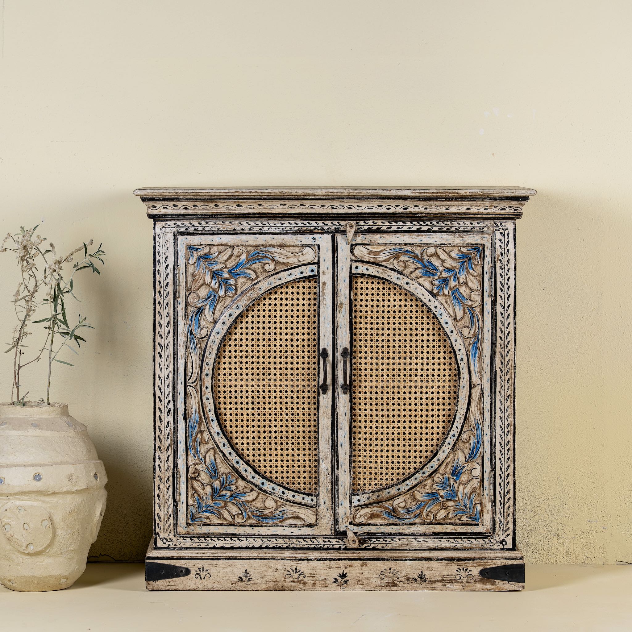 Living room scene of a hand-painted, hand-carved cupboard made of mango wood with floral design and rattan inserts.