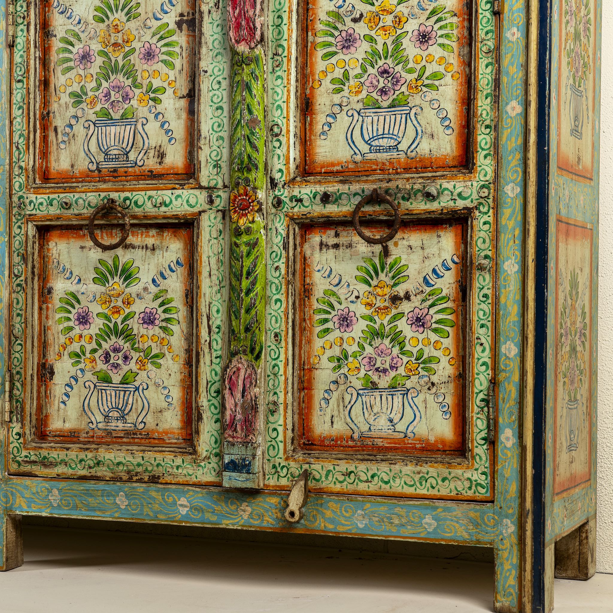 Close up image of the leg part of a hand-painted sideboard made from reclaimed wood, showcasing vibrant floral motifs in green, pink, yellow, and blue on a distressed turquoise base, with six panelled doors and rustic iron ring handles.