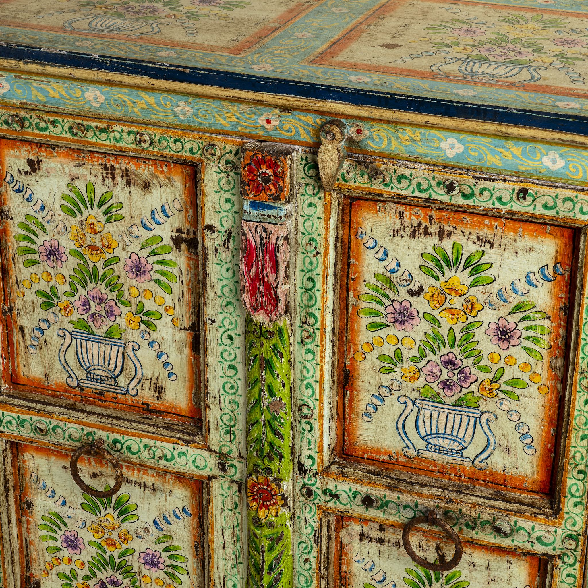 Close up image of a hand-painted sideboard made from reclaimed wood, showcasing vibrant floral motifs in green, pink, yellow, and blue on a distressed turquoise base, with six panelled doors and rustic iron ring handles.
