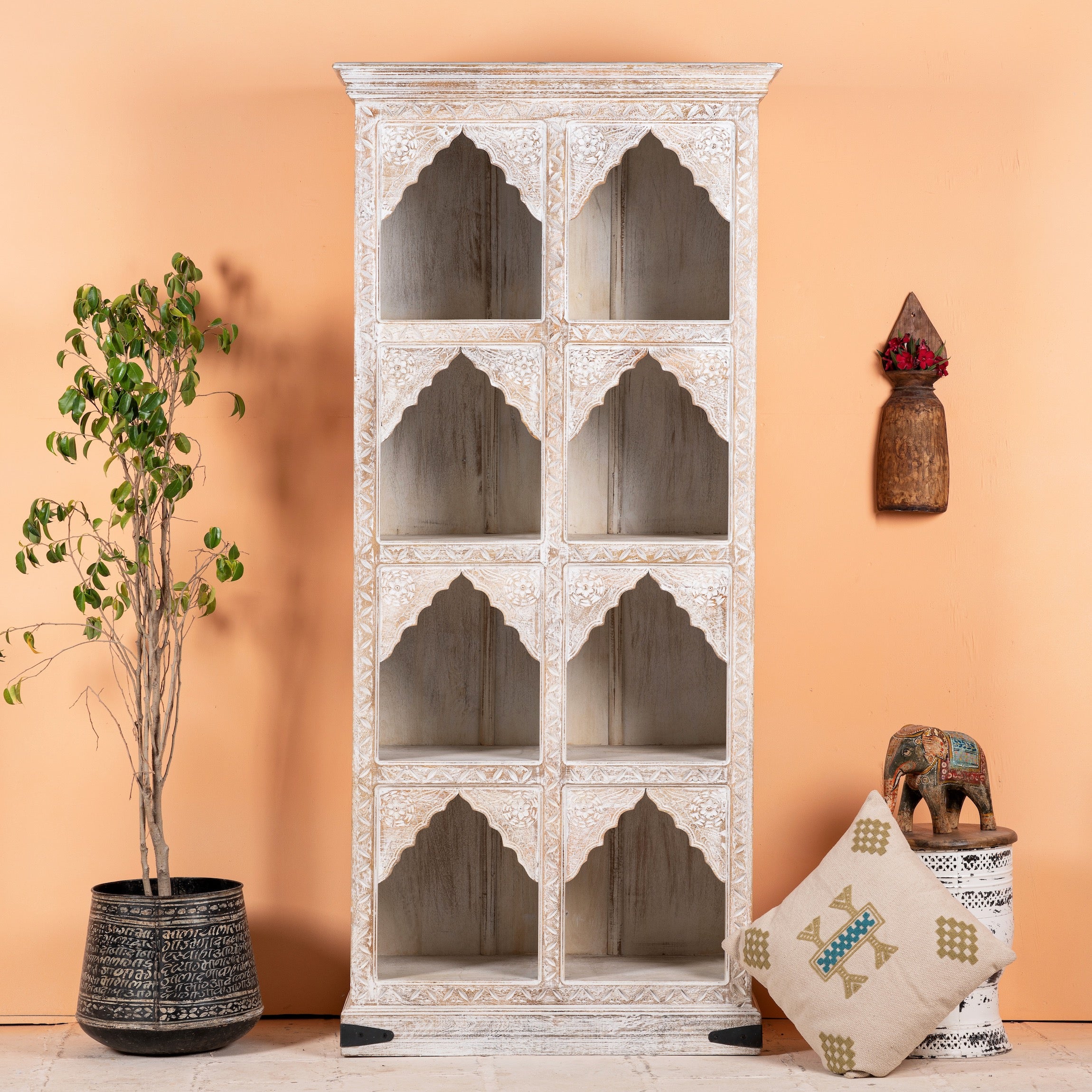 Hand-carved mango wood shelf with a whitewashed finish and Moroccan-style arches, featuring eight compartments, displayed in a home setting with decorative items and a potted plant beside it.