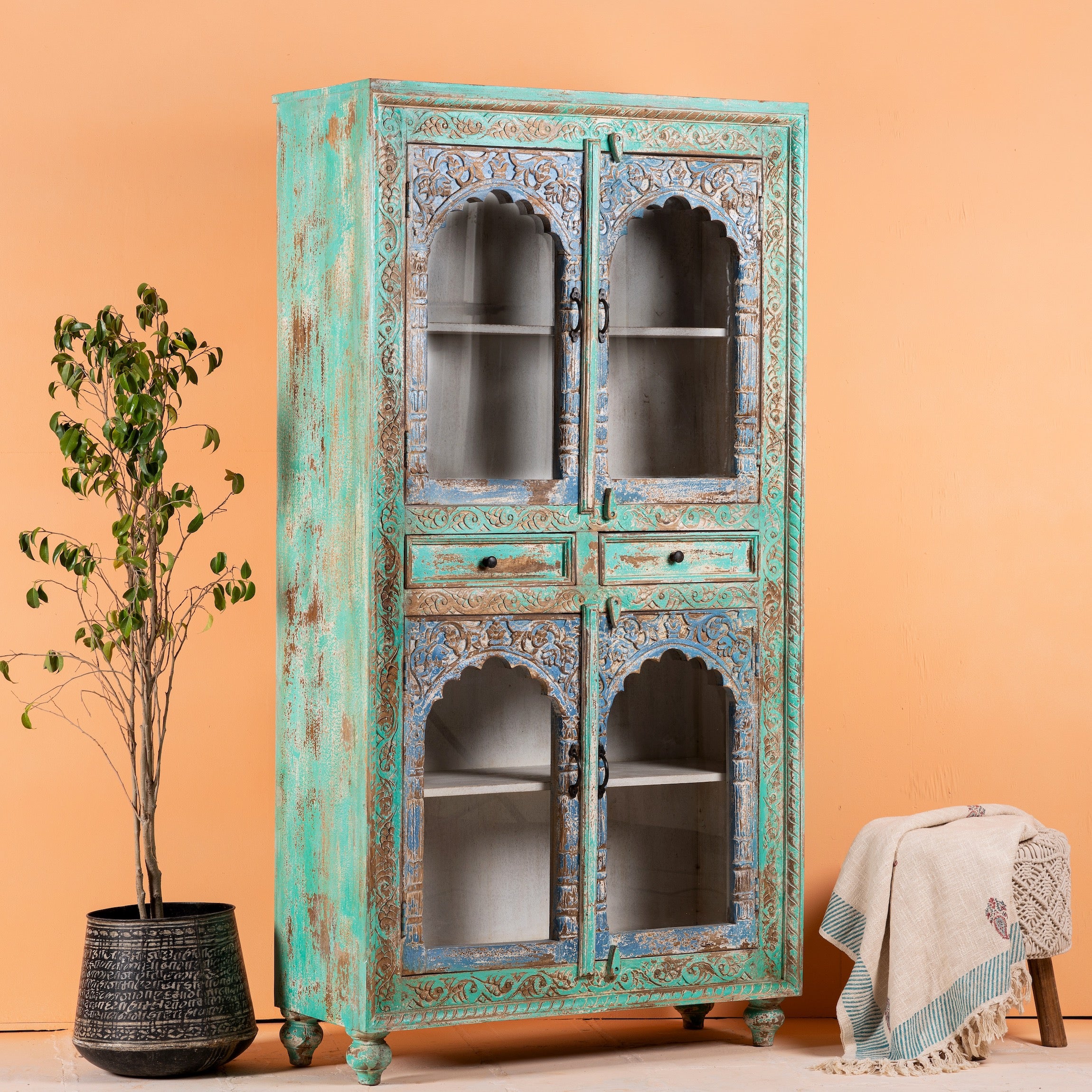 Side angle image of a hand-carved mango wood showcase with glass doors and drawers – carved wooden furniture in distressed turquoise and blue, combining artisan detail with vintage charm.
