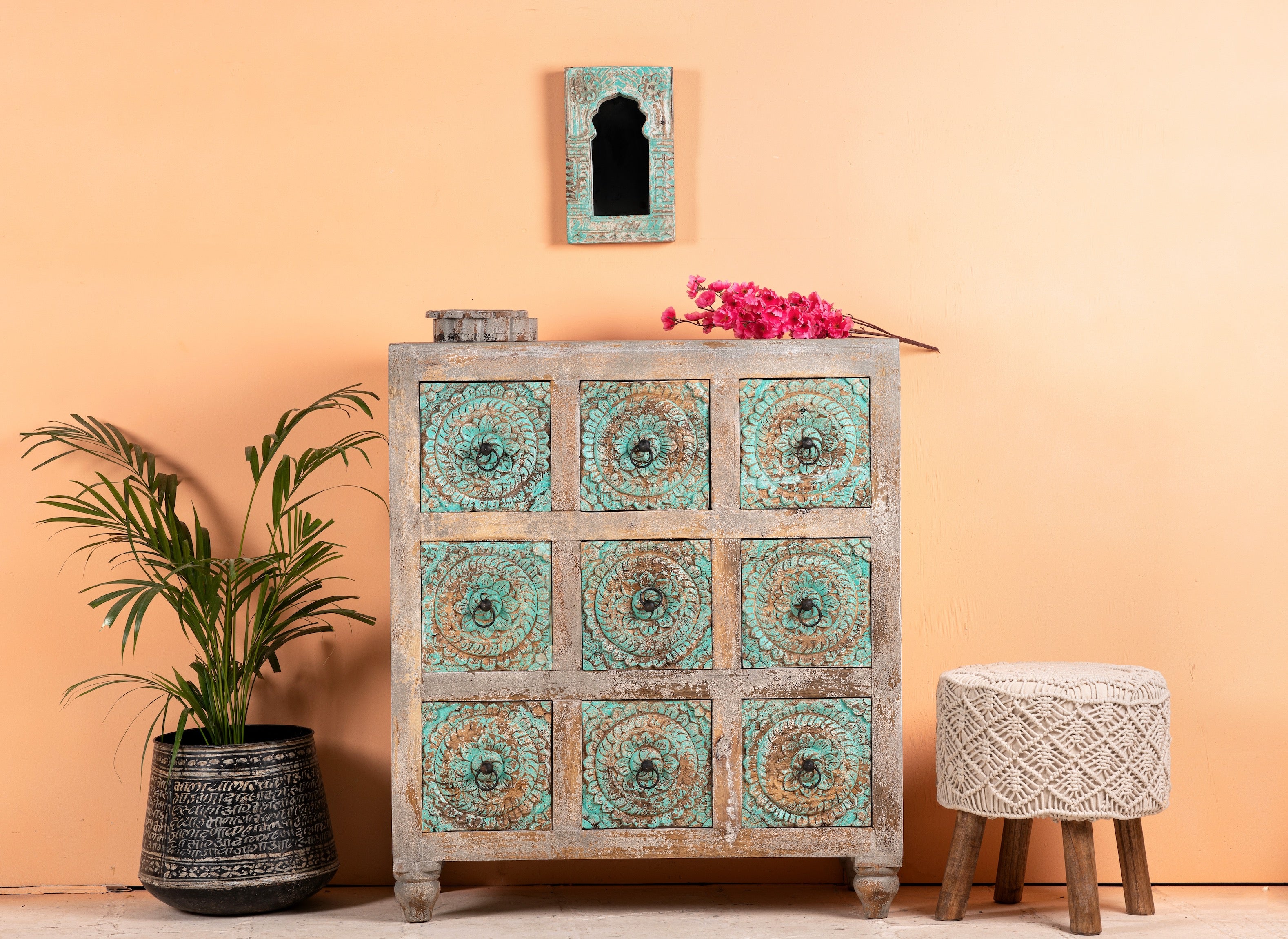 A vintage-style chest of nine drawers with hand-carved floral patterns on the drawer fronts, solid mango wood construction, and greenwashed detailing, placed against a warm-toned wall with a potted plant and decorative item on either side.