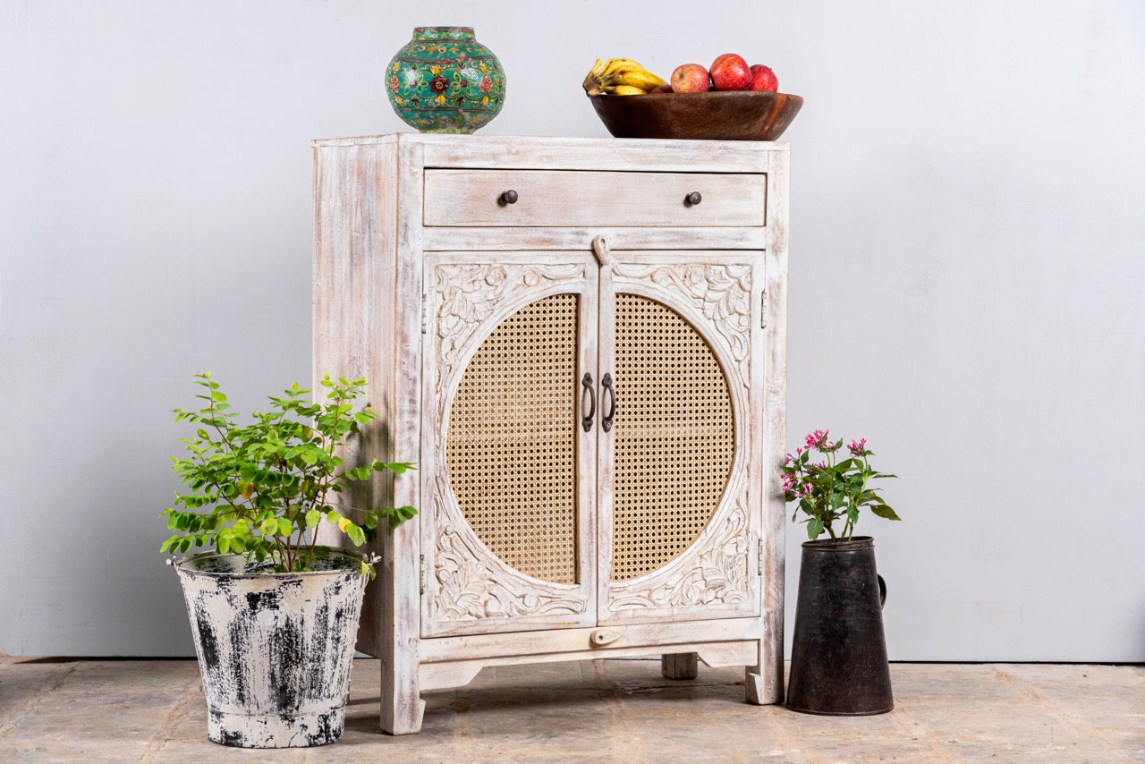 Side angle image of a white‑washed mango wood cupboard with rattan door panels and hand‑carved Indian ornamentation, 80 × 40 × 104 cm.
