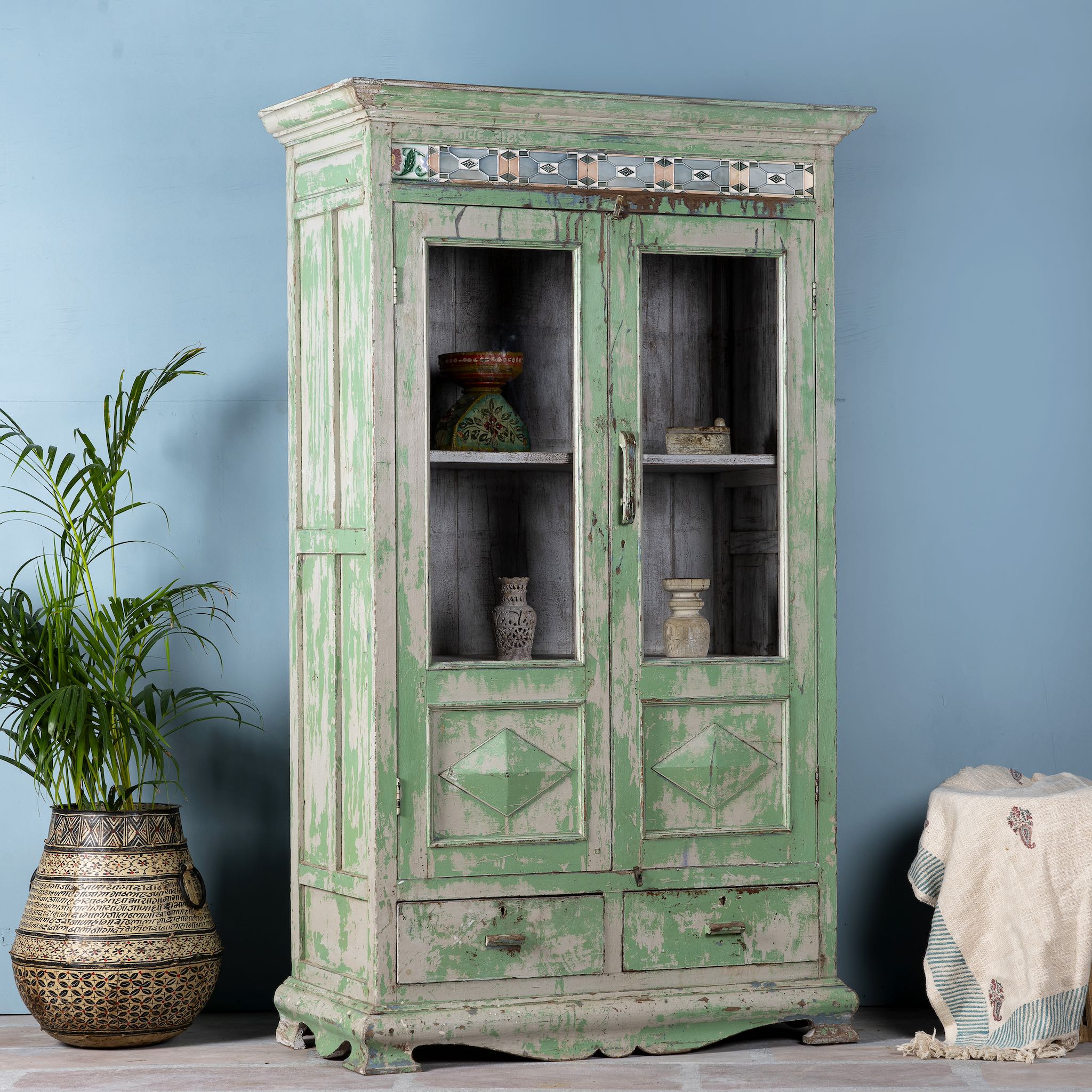 Side angle image of a reclaimed teak wood vitrine with distressed green finish, ceramic inlays, and glass-panelled doors – sustainable furniture with vintage Indian charm.