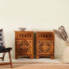 A pair of hand carved mango wood bedside tables with intricate floral and geometric carvings, finished in a warm teak colour, displayed in a room setting with decorative items on top.