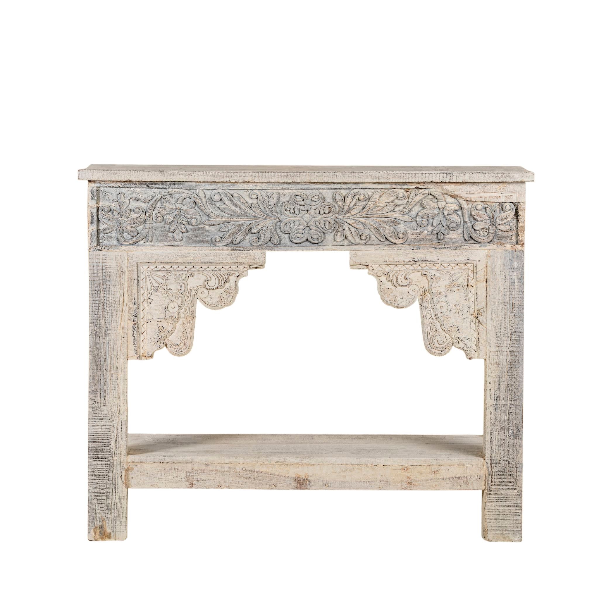 White background image of a refurbished teak wood console table with a whitewashed finish, featuring intricate hand-carved floral and scroll detailing on the front apron and corners, and a lower shelf for added display or storage.
