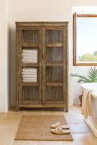 Lifestyle image of a handcrafted reclaimed teak wood display cabinet with glass doors and hand-painted Sanskrit symbols – sustainable furniture with vintage soul and artisanal charm.
