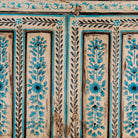 Close up image of a tall cabinet made from reclaimed wood, featuring hand-painted floral motifs in soft blue tones on panelled doors and drawers, with a distressed finish that adds vintage charm and highlights its artisanal craftsmanship.
