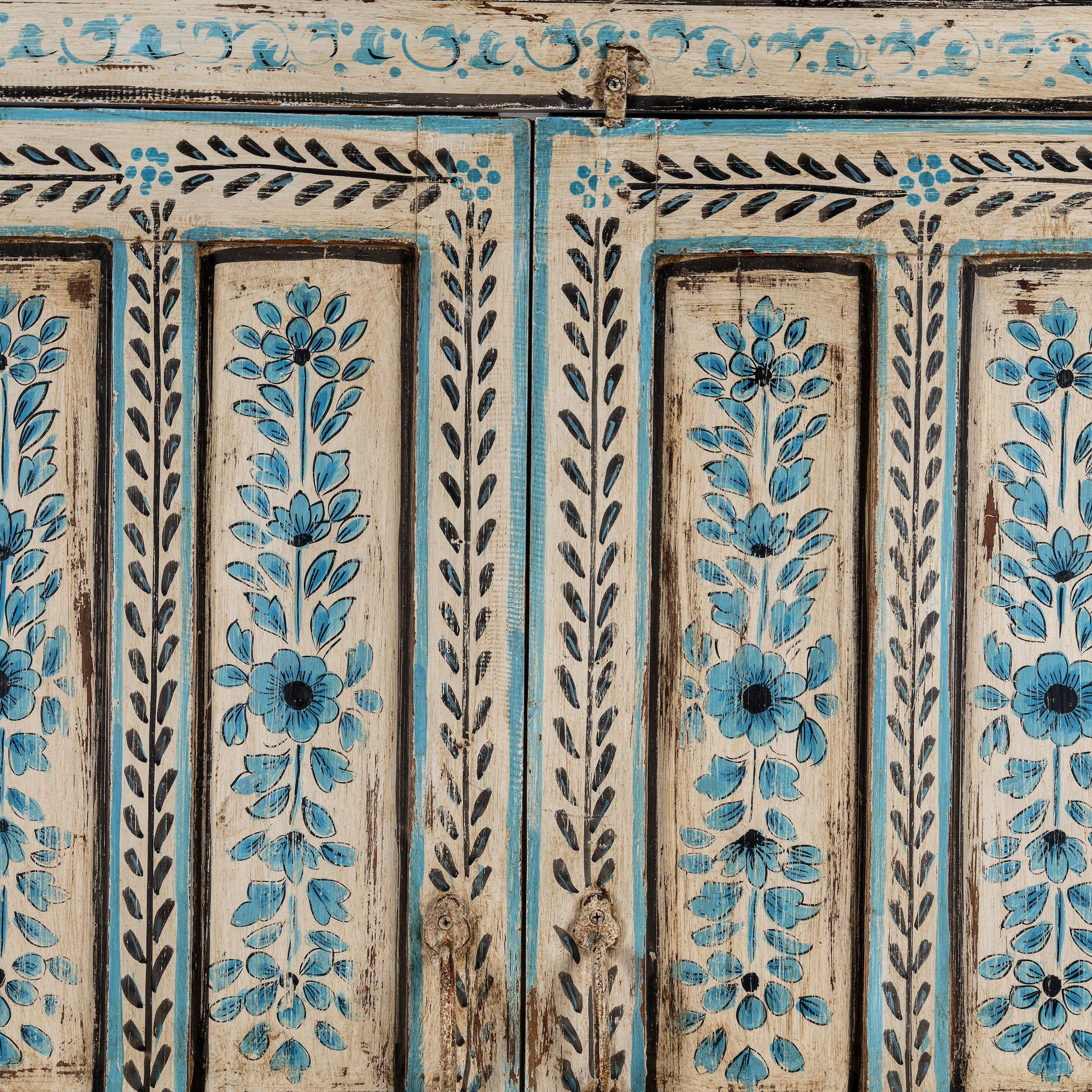 Close up image of a tall cabinet made from reclaimed wood, featuring hand-painted floral motifs in soft blue tones on panelled doors and drawers, with a distressed finish that adds vintage charm and highlights its artisanal craftsmanship.
