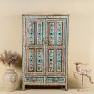 Front view image of a tall cabinet made from reclaimed wood, featuring hand-painted floral motifs in soft blue tones on panelled doors and drawers, with a distressed finish that adds vintage charm and highlights its artisanal craftsmanship.
