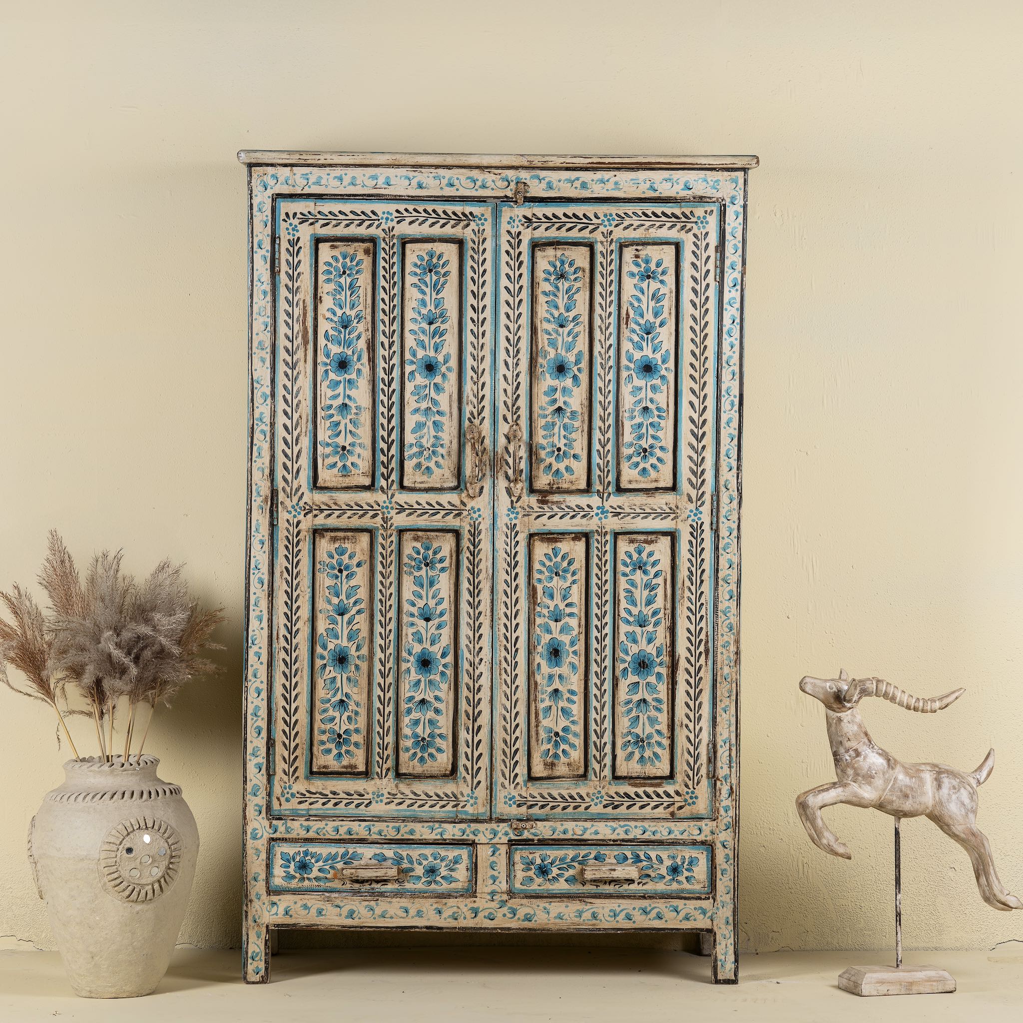 Front view image of a tall cabinet made from reclaimed wood, featuring hand-painted floral motifs in soft blue tones on panelled doors and drawers, with a distressed finish that adds vintage charm and highlights its artisanal craftsmanship.
