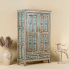 Side angle image of a tall cabinet made from reclaimed wood, featuring hand-painted floral motifs in soft blue tones on panelled doors and drawers, with a distressed finish that adds vintage charm and highlights its artisanal craftsmanship.
