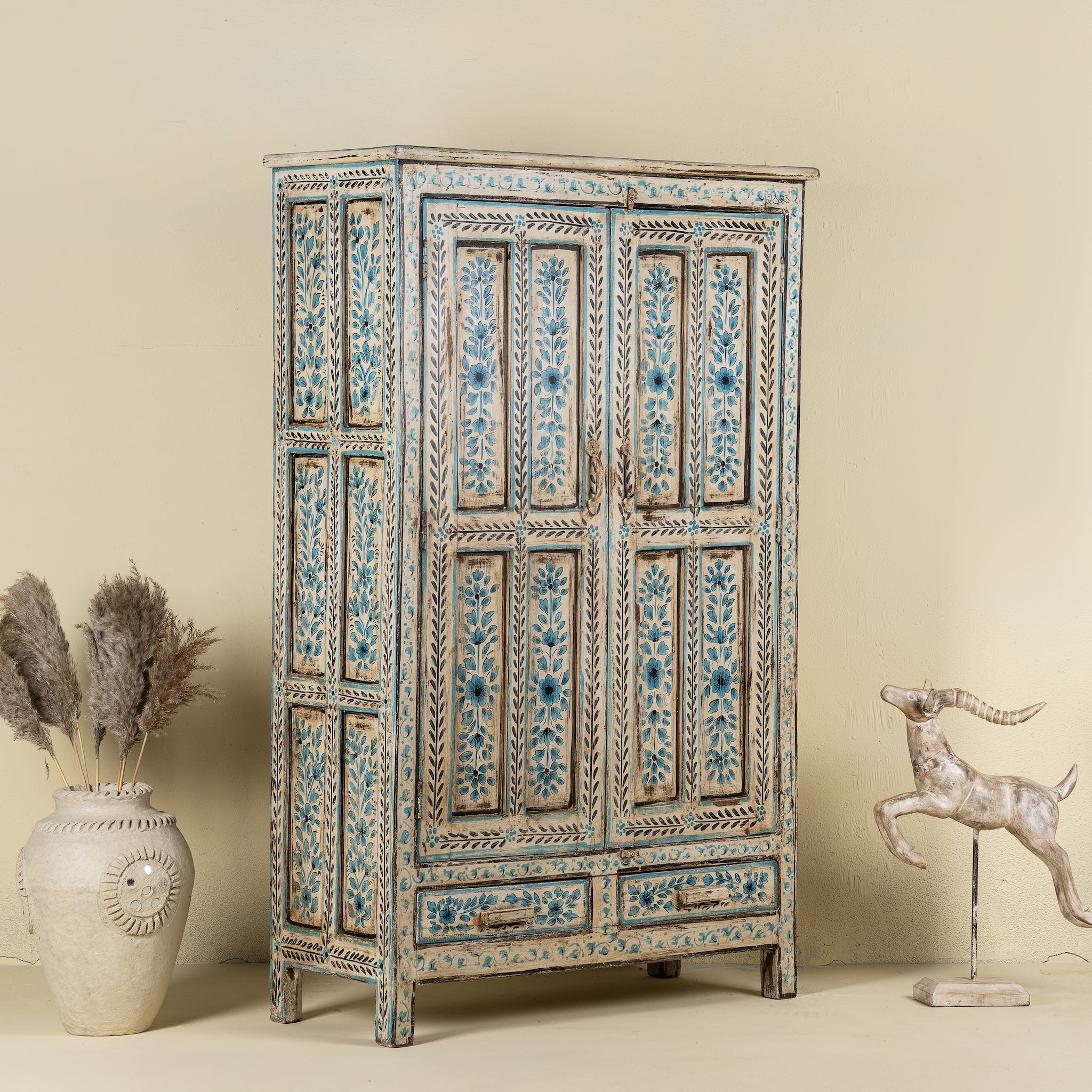 Side angle image of a tall cabinet made from reclaimed wood, featuring hand-painted floral motifs in soft blue tones on panelled doors and drawers, with a distressed finish that adds vintage charm and highlights its artisanal craftsmanship.
