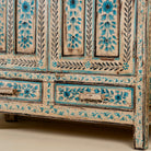 Close up image of the leg part of a tall cabinet made from reclaimed wood, featuring hand-painted floral motifs in soft blue tones on panelled doors and drawers, with a distressed finish that adds vintage charm and highlights its artisanal craftsmanship.
