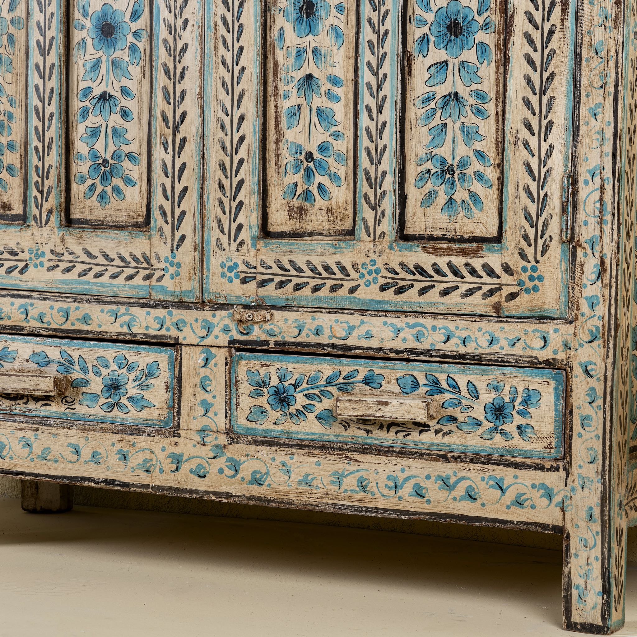 Close up image of the leg part of a tall cabinet made from reclaimed wood, featuring hand-painted floral motifs in soft blue tones on panelled doors and drawers, with a distressed finish that adds vintage charm and highlights its artisanal craftsmanship.
