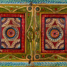 Close up image of a small hand-painted console made from reclaimed wood, featuring vibrant decorative motifs in red, green, blue, and gold on double doors, with intricate floral and geometric patterns and a lower open shelf for added display.

