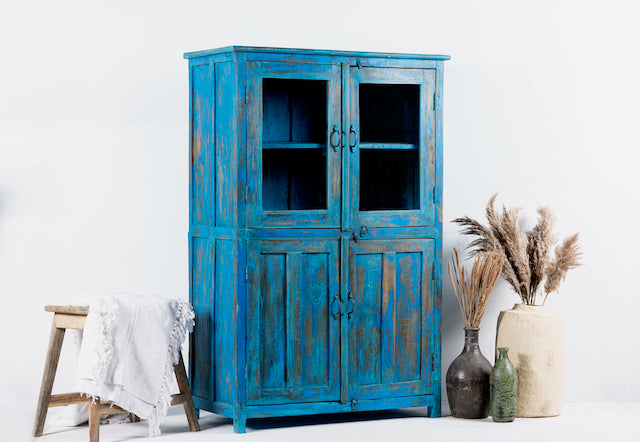 A vintage style refurbished teak wood showcase with blue paint and glass doors, accompanied by decorative items like a bamboo basket and a ceramic vase.