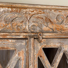 Close up image of a handcrafted showcase cabinet made from reclaimed wood, featuring diamond-pattern glass pane doors, a crackled paint finish in earthy tones, two drawers, and vintage-inspired detailing.