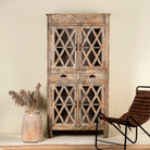 Handcrafted mango wood showcase cabinet with a rustic design, featuring glass-paneled doors and intricate wood carvings, placed in a room with a chair and decorative vase.