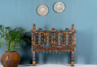 An antique teak wood Indian treasure chest with metal fittings and mirror inlays, featuring a colorful and ornate design, displayed against a blue wall with a plant and decorative plates above it.