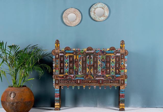 An antique teak wood Indian treasure chest with metal fittings and mirror inlays, featuring a colorful and ornate design, displayed against a blue wall with a plant and decorative plates above it.