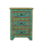 White background image of a  hand-painted reclaimed wood nightstand with vibrant colours and floral patterns, featuring three drawers, placed next to a vase with dried plants.