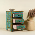 Open drawer image of a hand-painted reclaimed wood nightstand with vibrant colours and floral patterns, featuring three drawers, placed next to a vase with dried plants.